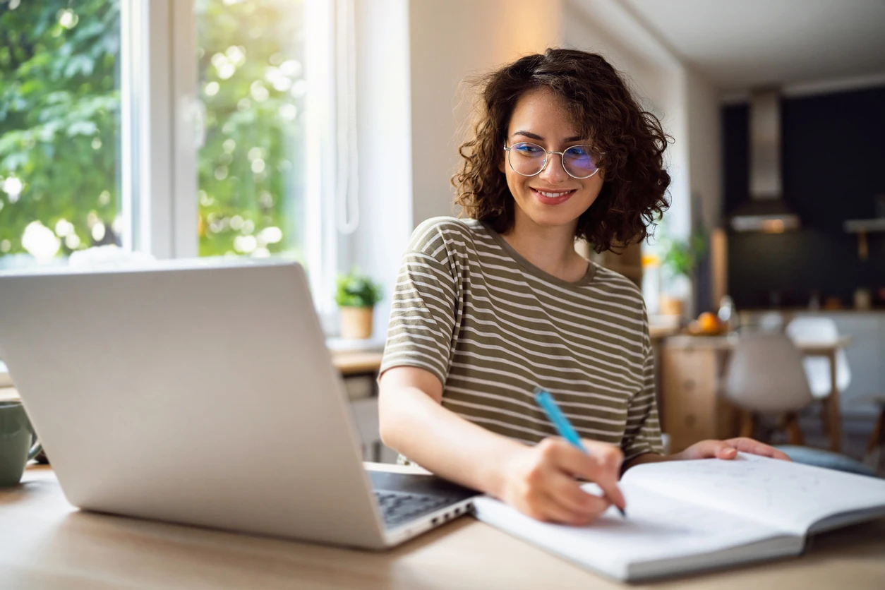 Student smiling while participating in online learning, taking notes in a notebook