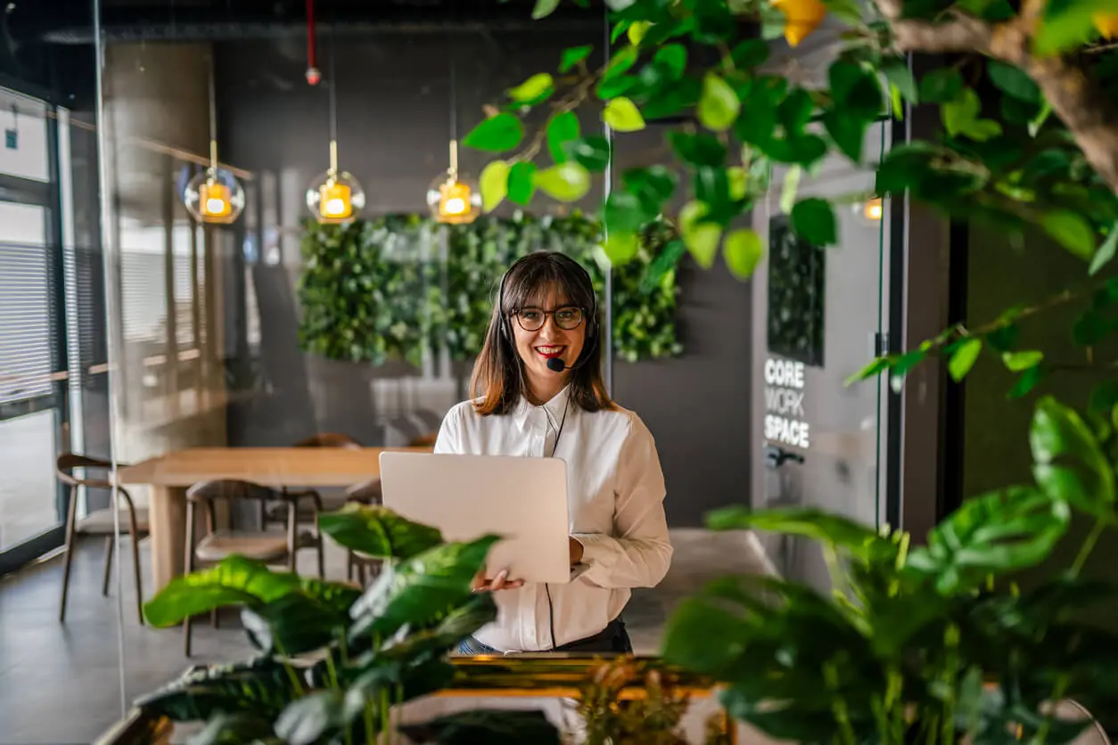 Person in a modern office with laptop and headphones, surrounded by greenery and pendant lights